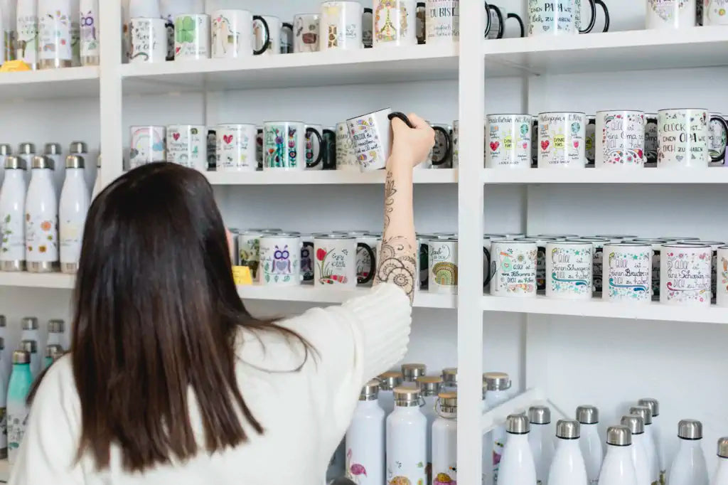 Tassen mit vielen verschiedenen Motiven in der Rollinart Galerie. Tina nimmt gerade eine Tasse in die Hand