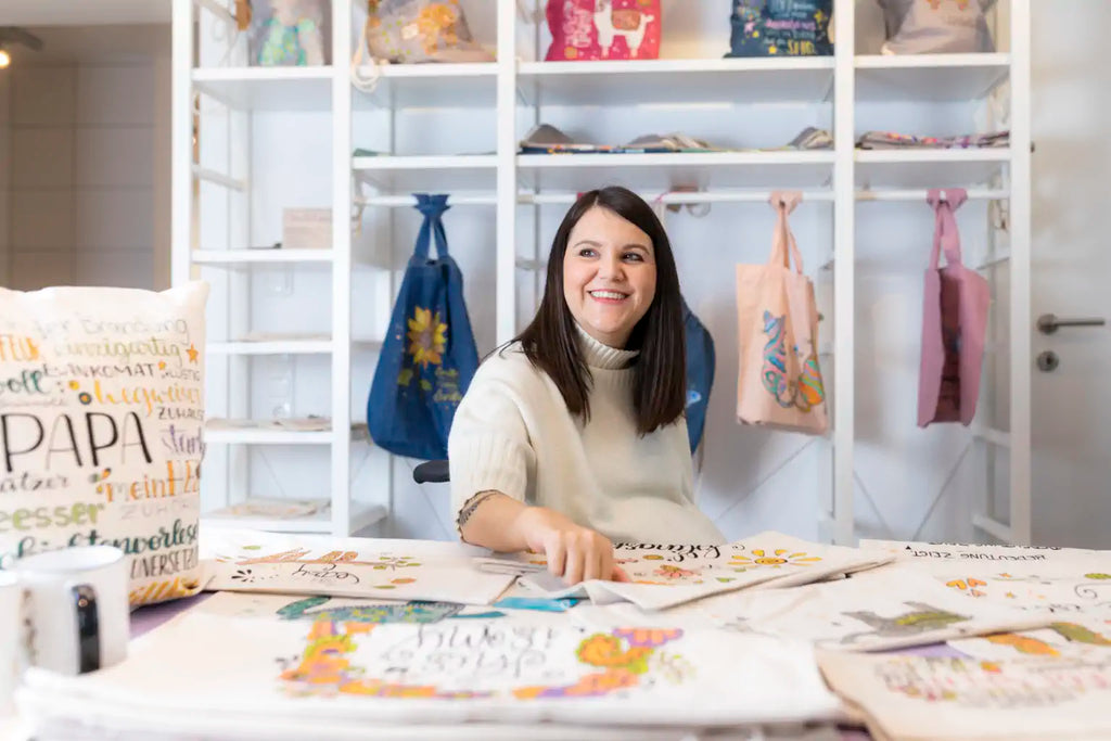Tina sitzt in der Galerie und lächelt. Man sieht Taschen und Textilien um sie herum.