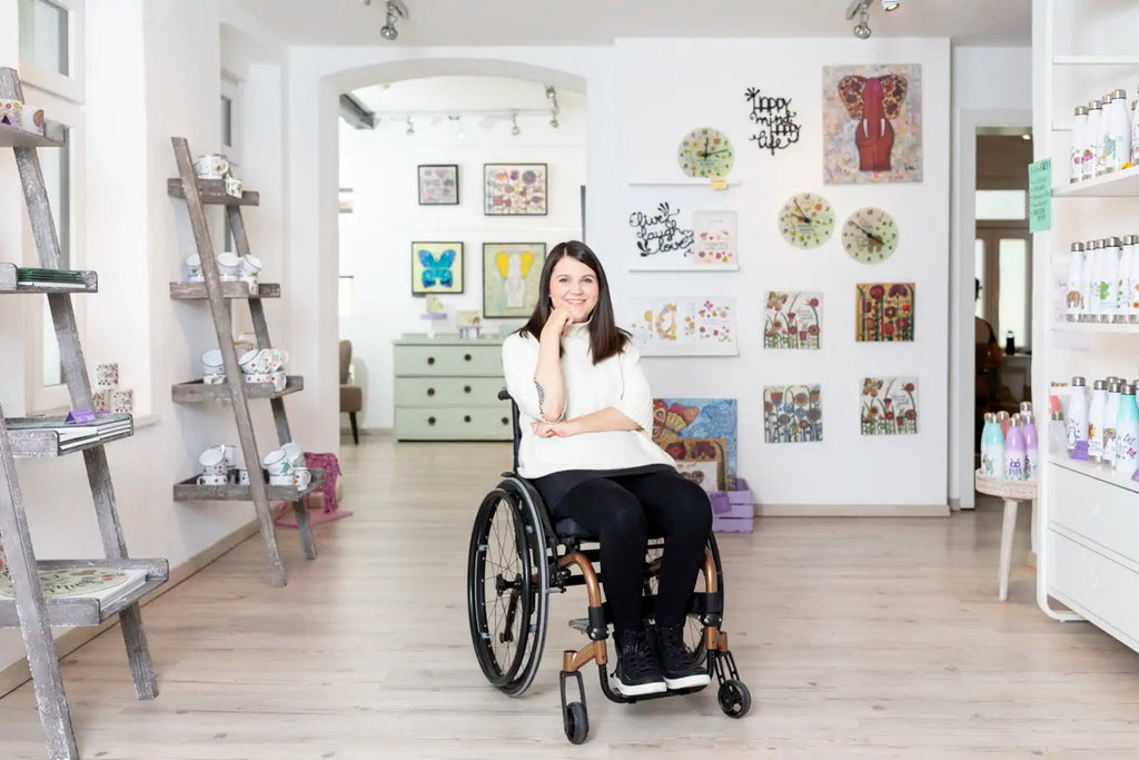 Tina sitzt im Rollstuhl in ihrer Galerie und lächelt freundlich in die Kamera