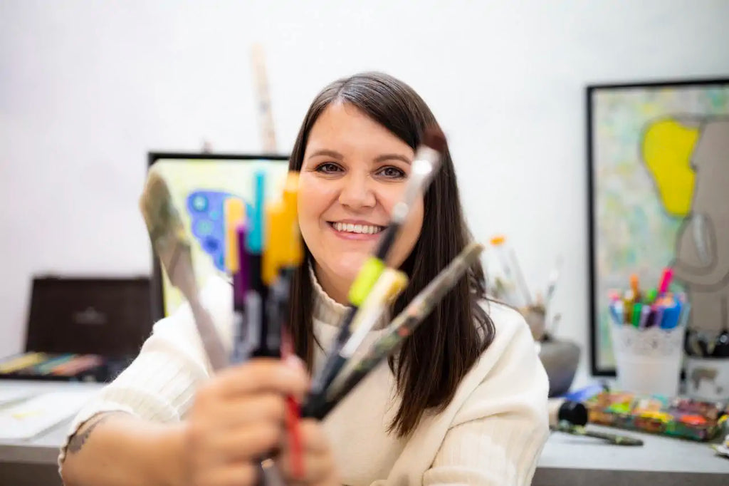 Tina hat viele Pinsel in der Hand und sitzt lächelnd vor 2 ihrer Bilder im Atelier